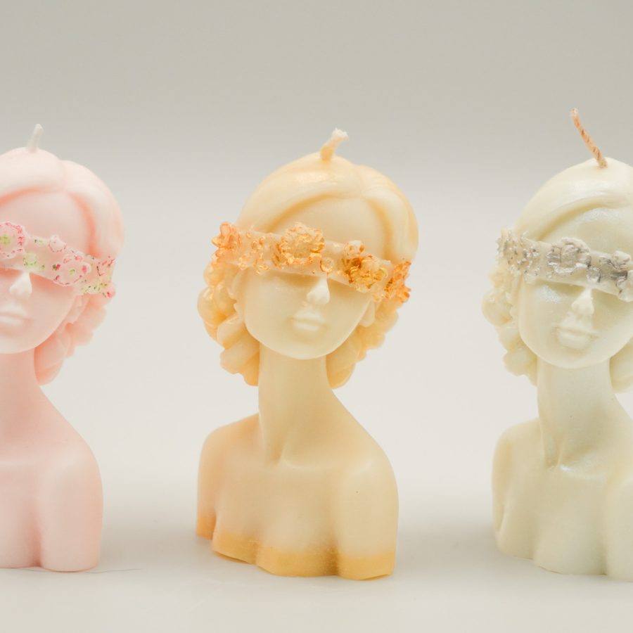 Three sculpted lady bust candles in pastel shades, each adorned with a detailed floral blindfold.