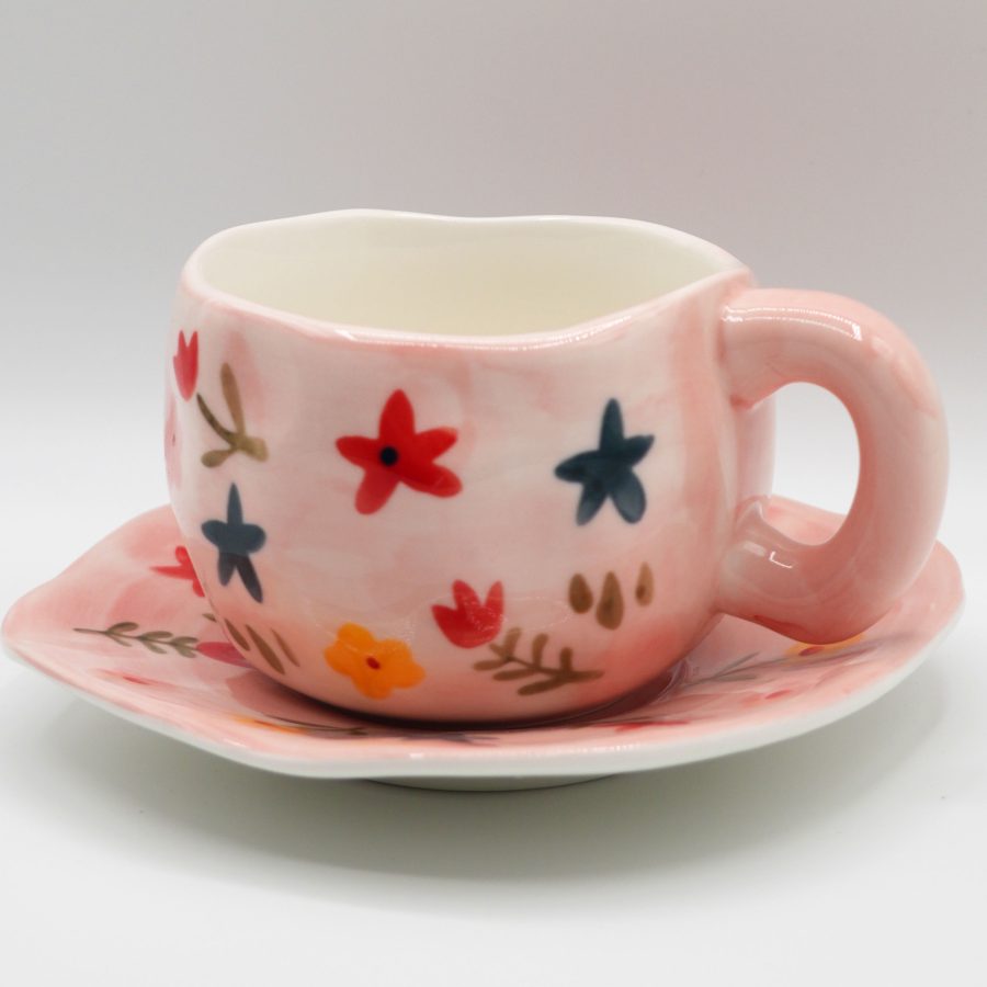 Chubby pink ceramic cup and matching saucer with colorful, folk-art style painted flowers.